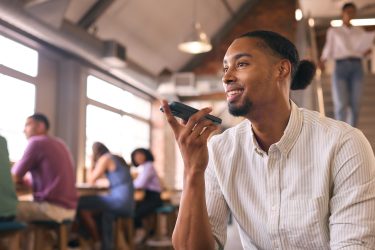 Businessman Talking Into Mic On Mobile Phone In Open Plan Office With Colleagues In Background